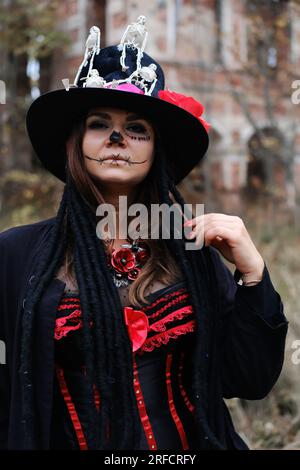 Eine Frau im Bild von Baron Saturday posiert vor dem Hintergrund eines alten Backsteingebäudes. Das Model trägt ein Korsett, eine Strickjacke und einen Hut Stockfoto