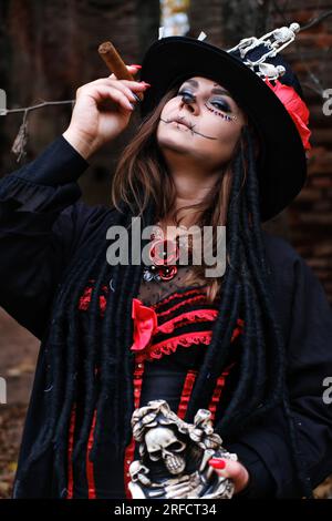 Eine Frau in Form von Baron Saturday mit einer Zigarre in der Hand. Das Modell trägt ein Korsett, eine Strickjacke und einen Hut mit Skelettfeige Stockfoto