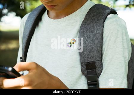 Teenager-Junge mit Autismus-Unendlichkeits-Regenbogen-Symbol mit Smartphone im Freien. Welttag des Autismus-Bewusstseins, Bewegung der Autismusrechte, Neurodiversität Stockfoto