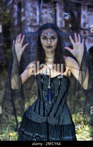 Eine geheimnisvolle gotische Frau in einem schwarzen Kleid mit einem Korsett, ihr Kopf ist mit einem schwarzen Schleier bedeckt. Model posiert vor einem alten verlassenen Stockfoto