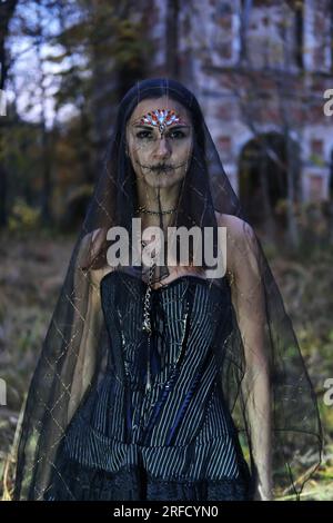 Eine mysteriöse Gothic-Frau in einem schwarzen Kleid mit einem Korsett, ihr Kopf ist mit einem schwarzen Schleier bedeckt. Model posiert vor einem alten verlassenen Gebäude Stockfoto