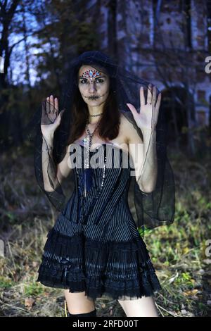 Eine mysteriöse Gothic-Frau in einem schwarzen Kleid mit einem Korsett, ihr Kopf ist mit einem schwarzen Schleier bedeckt. Model posiert vor einem alten verlassenen Gebäude Stockfoto