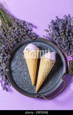 Zwei Lavendeleis in Waffle Cones auf einem Tablett und Blumensträuße aus trockenem Lavendel auf lila Hintergrund. Stockfoto