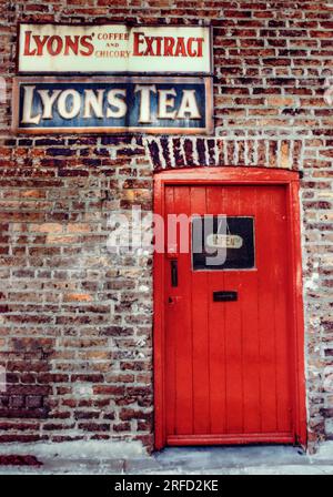 Village Shop außen im britischen Retro-Vintage-Charme mit „Open Sign“ im Türfenster. Charmanter, altweltenglischer Dorfladen mit roter Tür und verblassten Metallwerbeschildern für Lyons Coffee Extract & Lyons Tea an der umliegenden Backsteinmauer LACOCK WILTSHIRE UK Stockfoto