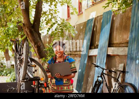 Enthusiastische, sportliebende afroamerikanische Frau mit Laptop, die Fahrrad im Hof repariert. Junge, sportliche schwarze Frau, die einen Minicomputer für die Fahrradwartung benutzt. Stockfoto