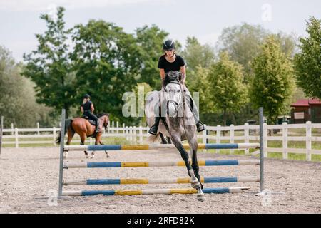 Eine Reiterin auf einem pappelgrauen Pferd, die an einem sonnigen Sommertag in der Outdoor-Arena über drei Bars springt. Pferdesport-Wettbewerbskonzept. Stockfoto