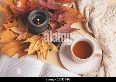 Tasse Tee auf einem Tisch, brennende Kerze mit Herbstblättern und offenem Buch. Gemütliches Zuhause, Herbstkonzept. Stockfoto