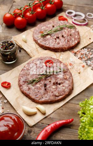 Rohe Burgerpasteten mit Gewürzen und Rosmarin auf Pergament, Sauce, frisches Gemüse, Hamburger-Zutaten auf Holzhintergrund. Stockfoto