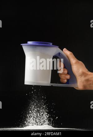 Die Hand einer Frau hält ein Öko-Plastiksieb auf schwarzem Hintergrund und siebt das Mehl. Küchenutensilien. Stockfoto