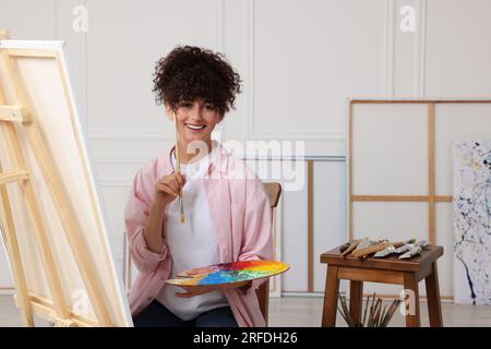 Junge Frau mit Pinsel und Künstlerpalette in der Nähe der Staffelei mit Leinwand Stockfoto