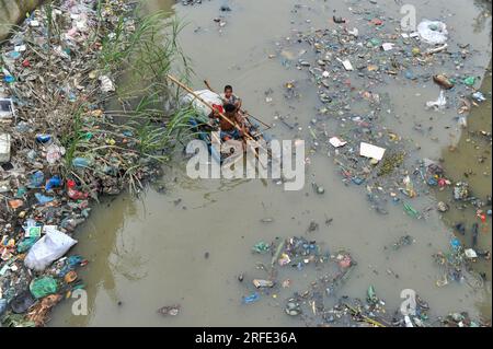 Ein Vater und Sohn rudern auf einem handgefertigten Floß und suchen nach Altkunststoff-Recyclingprodukten aus einem mit Kunststoffabfällen gefüllten Kanal im Kazir Bazar-Viertel von Sylhet. Dieser Kanal ist direkt mit dem Fluss Surma verbunden, der den Fluss Surma verschmutzt und das Flussbett füllt. Das Polyethylen hat sich in riesigen Haufen Schlamm angesammelt, was die Navigation behindert und zu einem Mangel an nutzbarem Wasser aus dem Surma-Fluss von Bangladesch führt. Sylhet, Bangladesch. Stockfoto