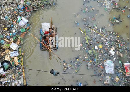 Ein Vater und Sohn rudern auf einem handgefertigten Floß und suchen nach Altkunststoff-Recyclingprodukten aus einem mit Kunststoffabfällen gefüllten Kanal im Kazir Bazar-Viertel von Sylhet. Dieser Kanal ist direkt mit dem Fluss Surma verbunden, der den Fluss Surma verschmutzt und das Flussbett füllt. Das Polyethylen hat sich in riesigen Haufen Schlamm angesammelt, was die Navigation behindert und zu einem Mangel an nutzbarem Wasser aus dem Surma-Fluss von Bangladesch führt. Sylhet, Bangladesch. Stockfoto