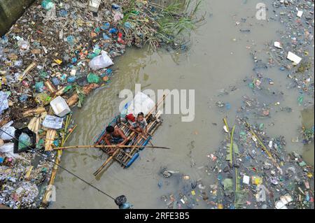 Ein Vater und Sohn rudern auf einem handgefertigten Floß und suchen nach Altkunststoff-Recyclingprodukten aus einem mit Kunststoffabfällen gefüllten Kanal im Kazir Bazar-Viertel von Sylhet. Dieser Kanal ist direkt mit dem Fluss Surma verbunden, der den Fluss Surma verschmutzt und das Flussbett füllt. Das Polyethylen hat sich in riesigen Haufen Schlamm angesammelt, was die Navigation behindert und zu einem Mangel an nutzbarem Wasser aus dem Surma-Fluss von Bangladesch führt. Sylhet, Bangladesch. Stockfoto