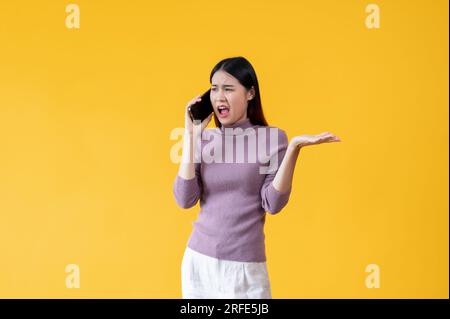 Eine wütende und unglückliche Asiatin spricht aggressiv am Telefon, beschwert sich am Telefon über jemanden, ist unzufrieden. Isoliertes Yello Stockfoto