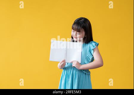 Ein hübsches kleines asiatisches Mädchen in einem blauen Kleid hält ein geöffnetes Buch mit leeren Seiten, während es auf einem isolierten gelben Hintergrund steht. Messwert, f Stockfoto