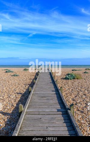 Promenade mit Bank am Dungeness Beach an der Südküste Großbritanniens. Kilometerlange einsame Kieselstrände, isolierte, schwarz bemalte Hütten und ein Kraftwerk! Stockfoto