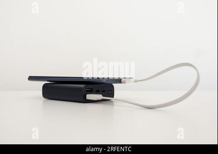 Aufladen des Mobiltelefons über die schwarze Powerbank auf dem Tisch. Weißer Hintergrund. Stockfoto