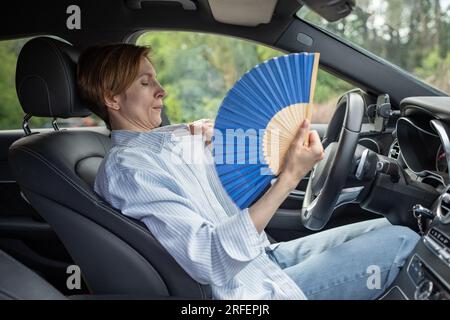 Eine müde, erschöpfte Frau mittleren Alters, die mit einem blauen Fächer wedelt, leidet an Steifigkeit im Auto. Stockfoto