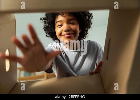 Ansicht von der Innenseite des Kartons. Afrikanisches Mädchen, das die Lieferung auspackt und in die Schachtel schaut. Frau öffnet Karton. Eine Frau bekommt ein Paket, das sich die gelieferten Waren ansieht. Zufriedenes positives Feedback des Kunden Stockfoto