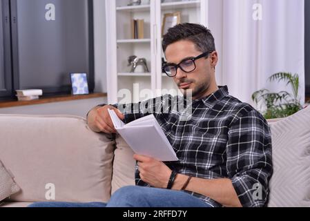 Intellektueller reifer Mann mit stilvoller Brille, in ein fesselndes Buch vertieft. Inmitten einer gemütlichen Einrichtung genießt er eine ruhige Lesesitzung auf dem Sofa des eigenen Zuhauses. Stockfoto