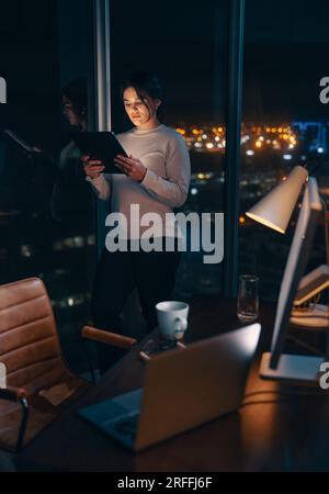 Engagierte kaukasische Geschäftsfrau, die bis spät in die Nacht in ihrem Büro arbeitet und ein digitales Tablet zur Datenanalyse verwendet. Unternehmerinnen lesen einen Bericht, m Stockfoto