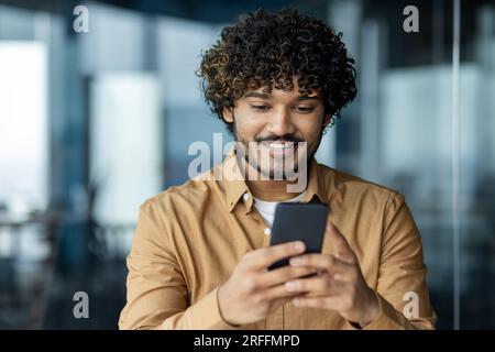 Nahaufnahme eines jungen Geschäftsmanns, der eine Online-Anwendung am Telefon verwendet, ein Mann im Büro, der gute Nachrichten vom Smartphone liest, ein lateinamerikanischer Mann, der zufrieden lächelt. Stockfoto