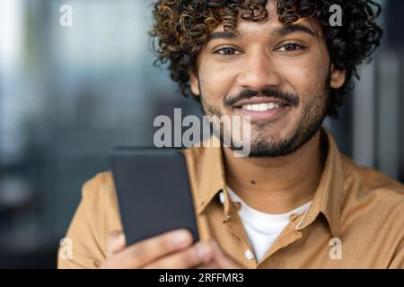 Nahaufnahme eines jungen hispanischen, glücklichen Geschäftsmanns, eines lächelnden Mannes, der in die Kamera schaut, eines Programmierers, der ein Smartphone in der Hand hält, der die App am Telefon verwendet. Stockfoto