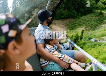 Vater und Kinder, die auf einem Sessellift in den Bergen fahren. Moderne Seilbahnhütten mit Menschen vor dem grünen Tal in den Bergen Stockfoto