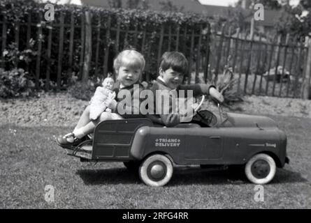 1950er Jahre, historisch, draußen in einem Hintergarten, ein kleiner Junge sitzt in seinem Tri-Ang Lieferwagen, einem mitfahrenden Spielzeugauto der Zeit, der seiner Schwester und ihrer Puppe eine Fahrt auf dem Rücken gibt, England, Großbritannien. Tri-Ang war ein führender Markenname des britischen Spielzeugautos Lines Bros in dieser Zeit, dem größten Spielzeugauto der Welt. Stockfoto