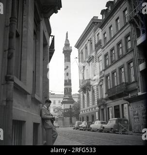 1950er, historisch, ein Mann mit einer Filmkamera steht in einer gepflasterten Seitenstraße mit Straßenbahnschienen in Paris, Frankreich, mit dem Kriegsdenkmal Colonne Vendome im Hintergrund. Ein verblichenes Poster für Le Cirque de Moscou an der Wand eines Gebäudes. Stockfoto