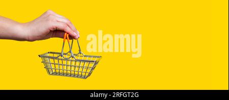 Die weibliche Hand hält den Warenkorb aus Spielzeug-Metall auf gelbem Hintergrund Stockfoto