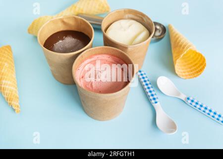 Eiscreme in einer handgefertigten Schachtel mit verschiedenen Geschmacksrichtungen – Erdbeeren, Schokolade, Vanille – steht auf blauem Hintergrund zusammen mit einem Waffeleisen und einem karierten Stockfoto