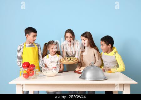 Weiblicher Koch mit zubereiteter Pizza und eine Gruppe kleiner Kinder während des Kochkurses auf blauem Hintergrund Stockfoto