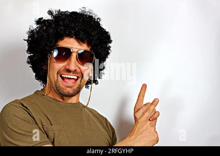 Positiv zufriedener junger Mann mit Afrohaar und Sonnenbrille, männlich sagt: Wow, wie aufregend es ist, hat einen erstaunlichen Gesichtsausdruck, deutet auf etwas hin. Auf weißem Backgr Stockfoto