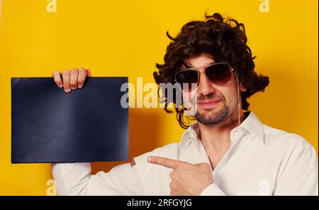Lockiger, bärtiger junger Mann, der lächelt und eine Sonnenbrille trägt und auf ein schwarzes, leeres Laken auf gelbem Hintergrund zeigt. Platz für Text . Studioaufnahme. Stockfoto