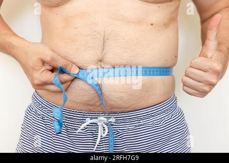 Der Mann misst seinen dicken Bauch mit Maßband und zeigt Daumen nach oben. Konzept der Gewichtsabnahme, Gesundheitsprobleme von übergewichtigen Menschen. Nahrungskontrolle und Akt Stockfoto