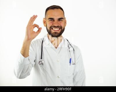 Junger männlicher Arzt, ok, unterschreiben, wenn er lächelt. Ich schaue auf die isolierte Kamera auf weißem Hintergrund Stockfoto
