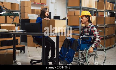 Männlicher Arbeiter mit Außerbetriebnahmen nimmt Kisten aus den Regalen im Lagerraum und organisiert Waren auf Regalen. Junger Mann im Rollstuhl, der mit einer Frau über Geschäftsentwicklung und Vertrieb spricht. Stockfoto