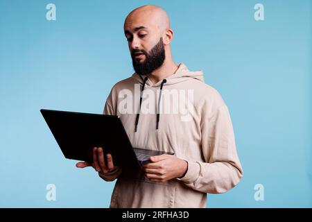 Ein seriöser arabischer Mann, der einen Laptop in der Hand hält und an einer Online-Videokonferenz teilnimmt. Junger Glatzkopf, der einen tragbaren Computer für Ferngespräche mit Web-Video-Software benutzt Stockfoto