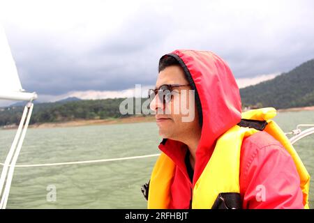 Lateinischer Erwachsener mit Sonnenbrille entspannt und glücklich mit Schwimmweste fährt ein Segelboot auf dem See, umgeben von Bergen Stockfoto