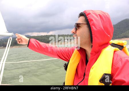 Lateinischer Erwachsener mit Sonnenbrille entspannt und glücklich mit Schwimmweste fährt ein Segelboot auf dem See, umgeben von Bergen Stockfoto