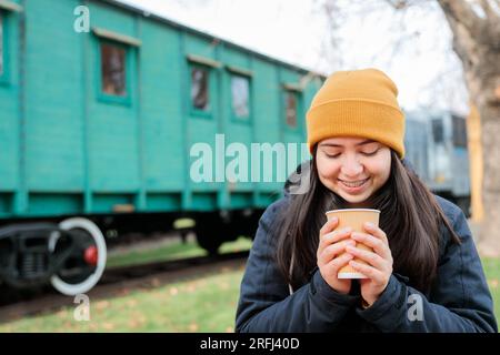 Kaffeepause im Bahnhofsgebäude: Junge Latina in warmer Kleidung, die eine Tasse Kaffee zum Mitnehmen isst. Speicherplatz kopieren Stockfoto