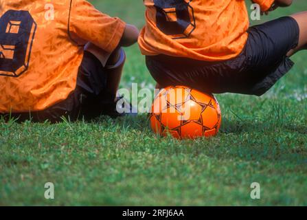 Ein Kinderfußballspieler sitzt auf einem Ball und sieht sich das Spiel an Stockfoto