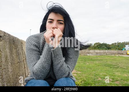 Porträt einer jungen kalten venezolanischen lateinamerikanischen Frau, die einen grauen Pullover trägt und Haare vom Wind weht, draußen sitzt und in die Kamera schaut, das Leben Stockfoto