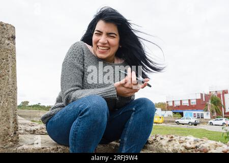 Eine junge latinatin, die glücklich am Telefon lacht, in sozialen Netzwerken surft, Nachrichten und Videos ansieht, draußen sitzt, sich ausruht, sehr unterhaltsam Stockfoto