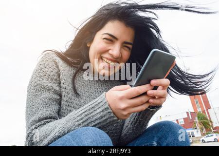 Eine junge latinatin, die glücklich am Telefon lacht, in sozialen Netzwerken surft, Nachrichten und Videos ansieht, draußen sitzt, sich ausruht, sehr unterhaltsam Stockfoto