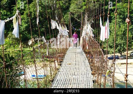 Ein bhutanischer Mann, der die traditionelle Gho trägt, geht über eine Hängebrücke im ländlichen Bhutan. Verblasste Gebetsfahnen hängen an den Metallseilen. Stockfoto