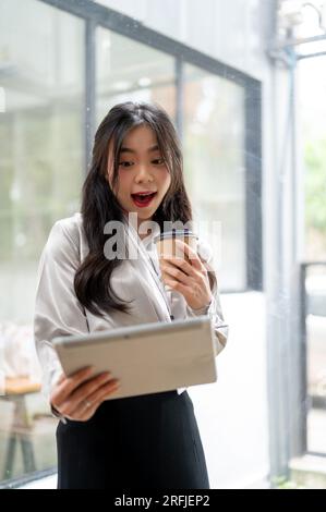 Ein Porträt einer schönen asiatischen Geschäftsfrau sieht mit einem aufgeregten und betäubten Gesicht auf ihren Tablet-Bildschirm, während sie in einem Büro-Corri Kaffee trinkt Stockfoto