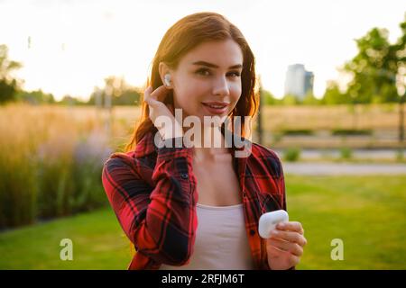 Eine lächelnde hübsche Frau steckt Kopfhörer in ihr Ohr, wenn sie auf der Straße steht. Hör der Musik zu. Stockfoto