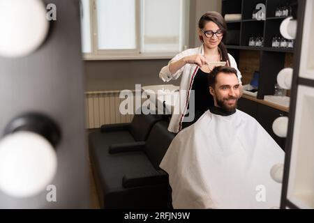 Handwerkerin in Gläsern schneidet Klienten Haare mit einer Schere unter dem Kamm Stockfoto